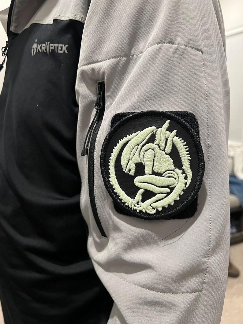 Op de afbeelding: Een zwart-grijze jas met een Kryptrek-logo en een ronde patch met een gloeiend groen alien-ontwerp. De jas heeft een zwart zakje met ritssluiting op de mouw.