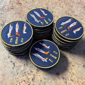 May include: Piles of circular navy blue embroidered patches with a gold border. Each patch displays three stylized aeroplanes in white and orange, with the words "FLY THEM ALL" in gold.