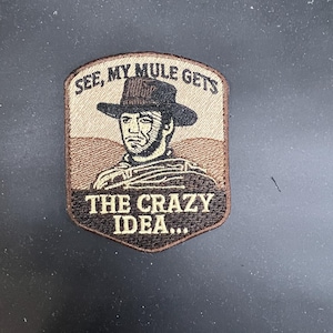 May include: Embroidered patch featuring a portrait of a man in a cowboy hat with the text "SEE, MY MULE GETS THE CRAZY IDEA...". The patch has a brown and tan color scheme, with a shield-like shape.