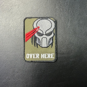 May include: A rectangular patch featuring the Predator's face with red laser sights and the text "OVER HERE" in white. The patch has a green background and a black border. The Predator's face is gray.
