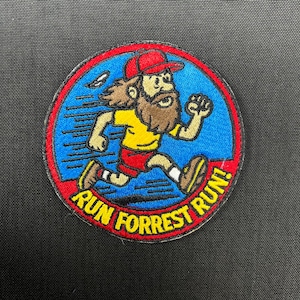 Puede incluir: Parche bordado con un personaje de dibujos animados corriendo. El personaje lleva una gorra roja, una camiseta amarilla y pantalones cortos rojos. El parche tiene un fondo azul con las palabras "RUN FORREST RUN!" en un borde rojo.