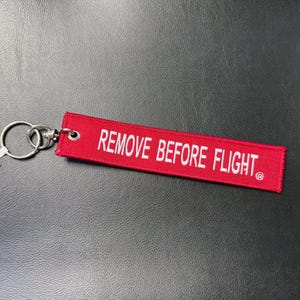 May include: A red keychain tag with the text "REMOVE BEFORE FLIGHT" in white. The tag is attached to a silver key ring and a small white tag. The background is a dark, textured surface.