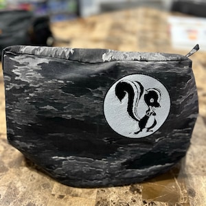 May include: A black and white zippered pouch with a camouflage pattern and a white circle with a black and white cartoon skunk in the center.