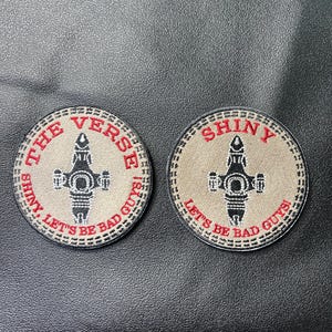 May include: Two circular patches with a tan background and a black spaceship design. The text "THE VERSE SHINY, LET'S BE BAD GUYS!" and "SHINY, LET'S BE BAD GUYS!" are in red around the edges. The patches are likely for clothing or accessories.