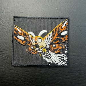 May include: Embroidered patch featuring a moth with orange, white, and yellow wings on a black square background. The moth has detailed wing patterns and a detailed body. The patch is likely for clothing or accessories.