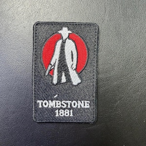 May include: Black rectangular patch featuring a stylized figure in a long coat and hat, holding a gun, set against a red circle. The text "TOMBSTONE 1881" is printed below the figure in white.