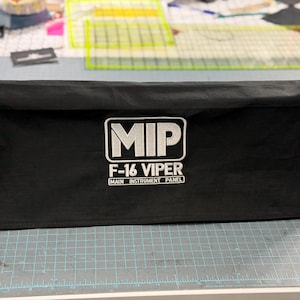 Può includere: Una copertura in tessuto nero di forma rettangolare con ricamo bianco che riporta la scritta MIP F-16 VIPER MAIN INSTRUMENT PANEL. La copertura presenta una struttura allungata con parte superiore piatta e lati dritti per proteggere un pannello.
