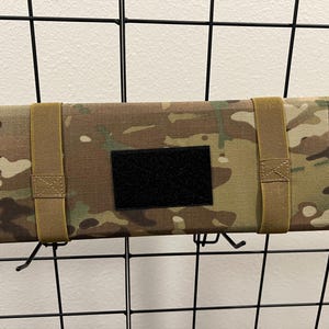 Könnte beinhalten: Eine Tasche in Tarnmuster mit zwei braunen Riemen und einem schwarzen Klettverschluss. Die Tasche hängt an einem schwarzen Metallgitter.