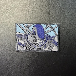 May include: Embroidered patch depicting an Alien creature. The patch shows the creature's head and upper body in blue, grey, and white against a light blue backdrop. The patch is rectangular in shape.