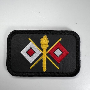 May include: Black leather patch with a gold and white embroidered design. Two crossed spears with red and white flags, and a gold torch in the center.