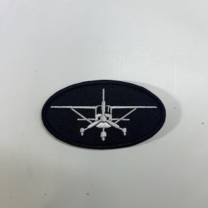 May include: Black oval patch featuring a white embroidered airplane design. The aircraft has a propeller and wings, set against a dark background. The patch is likely for aviation enthusiasts or as a decorative element.