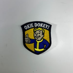 May include: Embroidered patch featuring a cartoon character giving a thumbs-up. The patch has a black border, a yellow background, and the words "OKIE DOKEY!" in white. The character is wearing a blue jumpsuit.