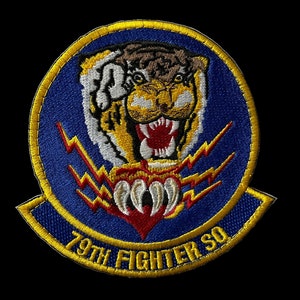 May include: A blue and yellow embroidered patch with a tiger's head and claws. The tiger is snarling with a red mouth and white teeth. The patch has the text "79th Fighter Sq" in yellow.