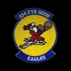 Op de afbeelding: Een geborduurde patch in blauw en geel met een rode adelaar die een voetbal draagt. De patch heeft de tekst "334 FTR SQDN EAGLES".