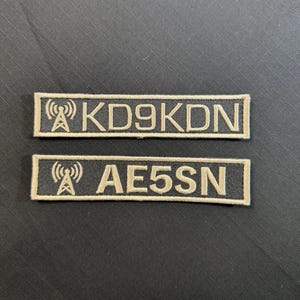 May include: Two rectangular embroidered patches with a dark gray background and light tan borders. Each patch features a radio tower icon on the left followed by alphanumeric text. The top patch reads KD9KDN and the bottom patch reads AE5SN.