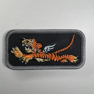 Może przedstawiać: Szara naszywka haftowana z czarnym tłem przedstawiająca pomarańczowego i czarnego tygrysa z skrzydłami. Tygrys biegnie i ma groźny wyraz twarzy.