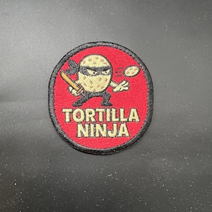 Peut inclure: Écusson brodé avec un fond rouge et une bordure noire. Le patch représente un personnage de ninja tortilla de dessin animé avec un masque noir et tenant une épée en bois. Le texte "TORTILLA NINJA" est en lettres blanches.