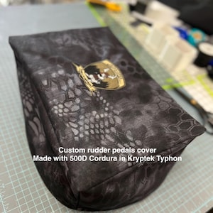 May include: Custom rudder pedals cover made with 500D Cordura in Kryptek Typhon. The cover is dark gray with a camouflage pattern and a gold and black emblem.