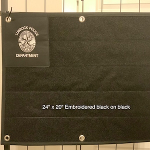 Puede incluir: Colgador de pared bordado negro, 61 cm x 51 cm, con el texto "Lubbock Police Department" y una insignia. El colgador tiene múltiples bolsillos horizontales y está montado en un marco de metal.