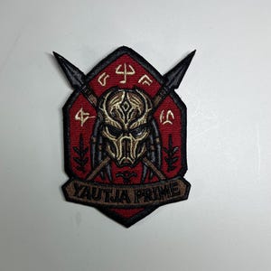 May include: Embroidered patch with a Predator mask design in gold and black, set against a red background. The patch includes the text "YAUTJA PRIME" in a banner, with two spears and symbols. The patch has a black border.