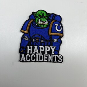 Puede incluir: Parche bordado que representa un orco de piel verde con armadura azul y detalles dorados. El parche dice "HAPPY ACCIDENTS" en letras blancas. El orco tiene una expresión de mueca.