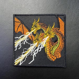 May include: Embroidered patch featuring a three-headed dragon with orange and yellow scales, wings, and lightning bolts against a black background. The patch is square-shaped and has a stitched border.