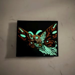 May include: A square canvas art piece featuring a moth design. The moth has orange and brown wings with white spots, and a glowing green body and accents. The background is black, and the artwork is displayed against a white wall.