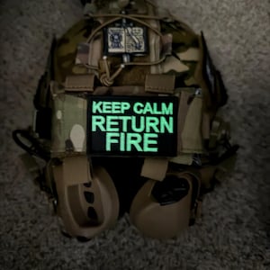 Może przedstawiać: Kamizelka taktyczna w kamuflażu z naszywką z napisem "KEEP CALM RETURN FIRE" w świecących na zielono literach. Kamizelka posiada różne kieszenie i mocowania, w zestawie ze słuchawkami.