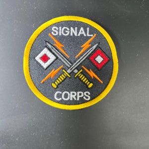 May include: Embroidered patch with a black background, yellow border, and the words "SIGNAL CORPS". Features crossed swords, lightning bolts, and signal flags. A military-themed patch.