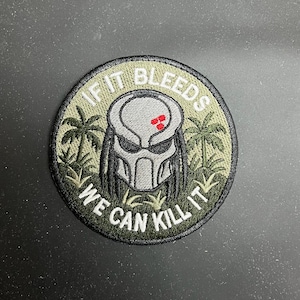 May include: Round embroidered patch featuring a Predator mask design in gray and black, with red accents. The patch is surrounded by green foliage and the text "IF IT BLEEDS WE CAN KILL IT" in white.