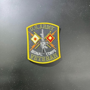Könnte beinhalten: Gestickter Aufnäher mit dem Text "U.S. ARMY SIGNAL CORPS VETERAN" in Gelb. Der Aufnäher hat einen schwarzen Hintergrund und einen gelben Rand. Er zeigt ein Gewehr und zwei rautenförmige Flaggen.