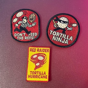 Könnte beinhalten: Drei gestickte Aufnäher. Ein roter ovaler Aufnäher mit einem Schiedsrichter-Motiv und dem Text DON'T FEED THE REFS!. Ein runder roter Aufnäher mit einer maskierten Tortilla-Figur und dem Text TORTILLA NINJA. Ein gelber rechteckiger Aufnäher zeigt eine rote Tornado-Grafik und den Text RED RAIDER TORTILLA HURRICANE.