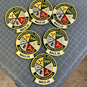 May include: Six yellow and black embroidered patches with the text "Nondestructive Inspection" and "What's Crackin?" with a logo of a yellow and black radiation symbol, a green and black waveform, a gray and black ruler, a red and black hand, and a gray and black water drop. The text "MALS-26" is also on each patch.