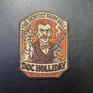 May include: Embroidered patch featuring a vintage illustration of a mustached man in a suit holding money and a revolver. The text reads "THE DENTIST FROM HELL" and "DOC HOLLIDAY" in a stylized font, with orange and brown colors.