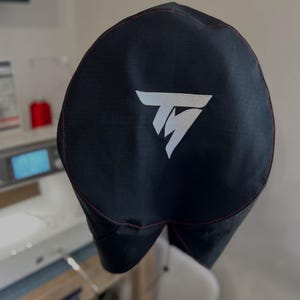 May include: Black fabric cover with a white logo, featuring a stylized "TM" design. The cover has red stitching around the edges. The background shows a sewing machine and a spool of red thread.