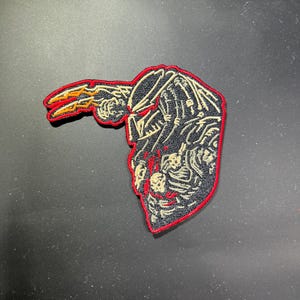 May include: Embroidered patch featuring a Predator design. The patch has a black background with a red border. The Predator is depicted with a black helmet, red eyes, and a beige face. The Predator's weapon is orange and yellow.