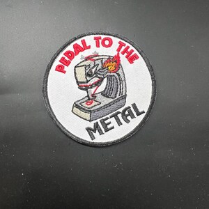 May include: Embroidered patch with the text "PEDAL TO THE METAL" in red, surrounding a stylized image of a machine with flames. The patch is circular, with a white background and a black border.