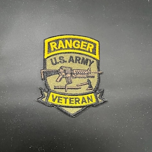 May include: Embroidered patch with a yellow border and text that reads "RANGER U.S. ARMY VETERAN". The patch features a rifle, a hatchet, and a knife. The background is olive green.