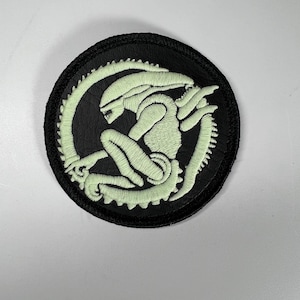 Puede incluir: Un parche redondo bordado en negro con un diseño de alienígena verde claro. El alienígena está en posición curvada, rodeado por una cola segmentada. El parche tiene un borde negro y es adecuado para ropa o accesorios.