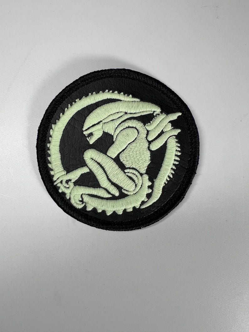 Op de afbeelding: Een ronde, zwart geborduurde patch met een lichtgroen alien-ontwerp. De alien is in een gekrulde positie afgebeeld, omringd door een gesegmenteerde staart. De patch heeft een zwarte rand en is geschikt voor kleding of accessoires.