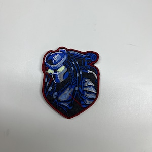 May include: Embroidered patch featuring a blue and black Predator design with a red border. The patch depicts the Predator's helmet and shoulder armor. The patch is likely for jackets, bags, or other apparel.