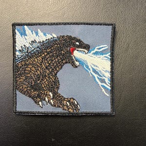 May include: Embroidered patch featuring Godzilla, a large, dark brown and black monster, exhaling a blue and white beam. The patch has a gray background and a black border.