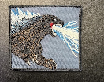 Op de afbeelding: Geborduurde patch met Godzilla, een groot, donkerbruin en zwart monster, dat een blauwe en witte straal uitstoot. De patch heeft een grijze achtergrond en een zwarte rand.