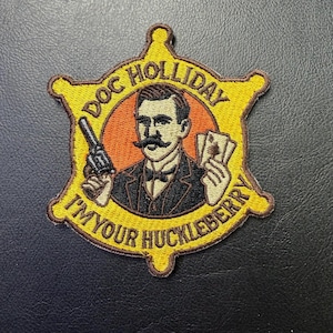 May include: Embroidered patch featuring Doc Holliday in a yellow sheriff badge shape. The patch includes the text "DOC HOLLIDAY" and "I'M YOUR HUCKLEBERRY". Doc Holliday is depicted with a mustache, holding a gun and playing cards.
