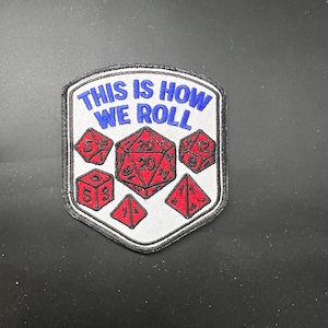 May include: A white patch with a black border featuring the text "THIS IS HOW WE ROLL" in blue. It also displays red dice with white numbers, including a D20, D6, D4, and D12, perfect for tabletop gaming enthusiasts.
