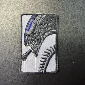 May include: Embroidered patch featuring a detailed depiction of an Alien creature. The design showcases the creature's head and part of its body in shades of black, gray, and blue, set against a light gray background. The patch has a rectangular shape with a black border.