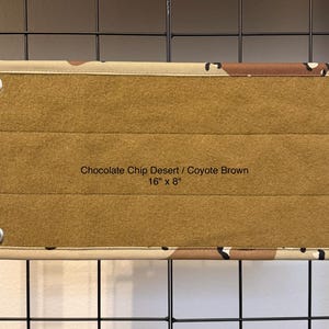 May include: A rectangular fabric panel featuring a chocolate chip desert camouflage border and a solid coyote brown center. The panel includes four silver metal grommets at the corners and is printed with the text Chocolate Chip Desert / Coyote Brown 16" x 8".