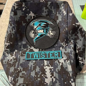 May include: A backpack with a digital camouflage pattern in shades of gray and black. It features a circular patch with a blue and white tornado graphic and a rectangular patch that reads "TWISTER" in teal.
