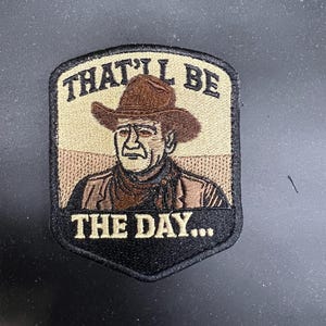 May include: Embroidered patch featuring a cowboy with a brown hat and vest. The patch has the text "THAT'LL BE THE DAY..." in white lettering. The patch is bordered in black and has a tan and brown background.