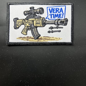 Op de afbeelding: Geborduurde patch met een gestileerd geweer met een vizier, in tinten beige, zwart en bruin. De tekst "VERA TIME!" staat in een blauwe tekstballon. De patch heeft een zwarte rand en een witte achtergrond.
